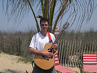 man holdling guitar with palm trea and beach dunes in background