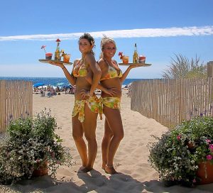 2 smiling servers in bikinis in the sand holding drink trays